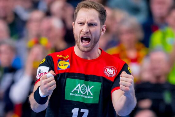 Ein männlicher Handballspieler, der ein schwarz-rotes Trikot mit der Nummer 17 trägt, jubelt mit geballten Fäusten und scheint während eines Spiels leidenschaftlich zu schreien, während im Hintergrund eine verschwommene Menschenmenge zu sehen ist.
