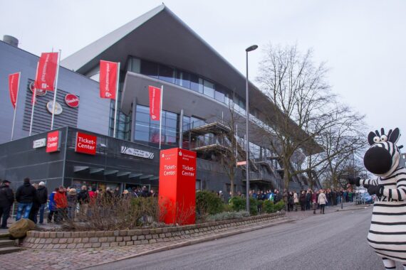 Menschen stehen vor einem modernen Gebäude mit roten Schildern mit der Aufschrift Ticket Center an. Eine Person in einem Zebrakostüm steht auf der Straße und winkt. Bäume und bedeckter Himmel sind zu sehen.