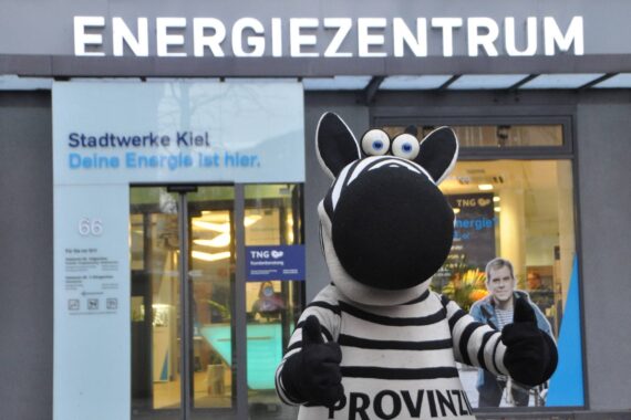 Eine Person im Zebrakostüm steht mit erhobenem Daumen vor dem Gebäude des Energiezentrums, hinter dem Glastüren und Schilder in deutscher Sprache zu sehen sind.
