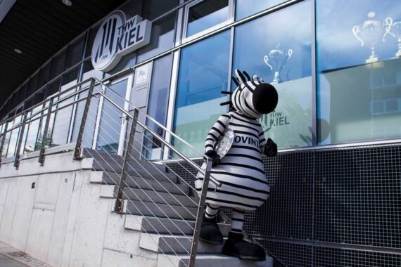 Eine Person in einem schwarz-weiß gestreiften Zebrakostüm steht auf einer Außentreppe in der Nähe eines modernen Gebäudes mit großen Fenstern und dem Logo "THW Kiel" über dem Eingang.