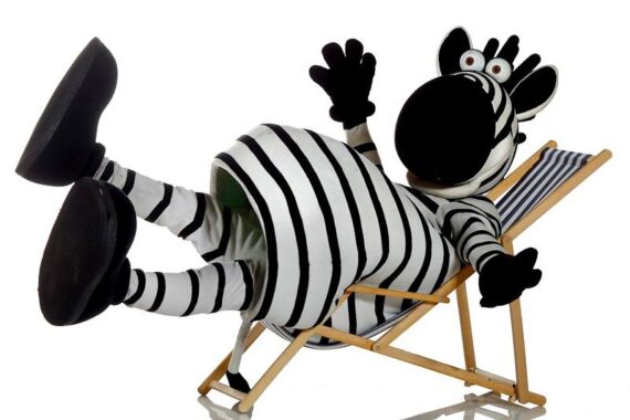 Eine Person in einem skurrilen Zebrakostüm lümmelt sich spielerisch auf einem gestreiften Liegestuhl, die Beine hochgelegt und einen Arm erhoben, vor einem schlichten weißen Hintergrund.