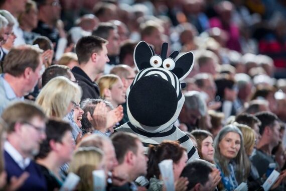 Eine Person in einem großen Zebrakostüm sitzt bei einer Indoor-Veranstaltung inmitten einer Menschenmenge und sticht unter den jubelnden und klatschenden Zuschauern hervor.