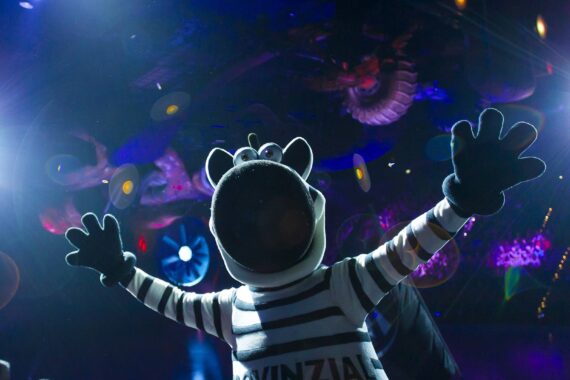 Eine Person in einem Zebra-Maskottchen-Kostüm steht mit weit ausgebreiteten Armen unter hellem Bühnenlicht, umgeben von farbenfrohen, skurrilen Dekorationen in einer lebhaften Innenumgebung.