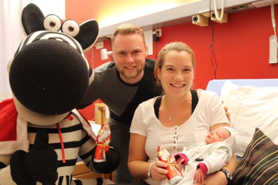 Eine lächelnde Mutter mit einem Neugeborenen, die neben einem Mann und einer Person in einem Zebra-Maskottchenkostüm sitzt. Die Erwachsenen halten jeweils eingepackte Schokoladenweihnachtsmänner in einem hell erleuchteten Krankenhauszimmer.
