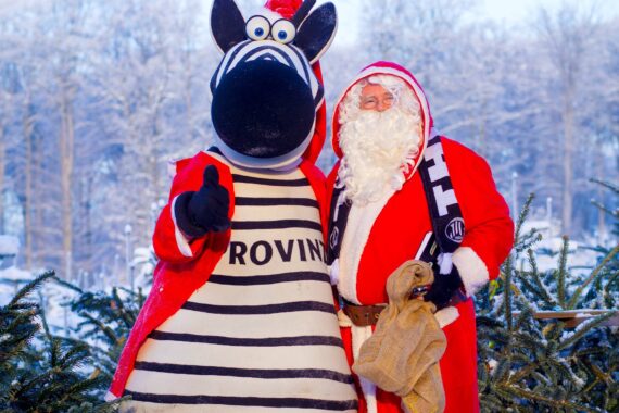 Eine Person in einem Weihnachtsmannkostüm steht neben einem großen Zebra-Maskottchen mit roter Jacke und gestreiftem Hemd. Sie lächeln und posieren im Freien vor verschneiten Bäumen und grünen Tannenzweigen.