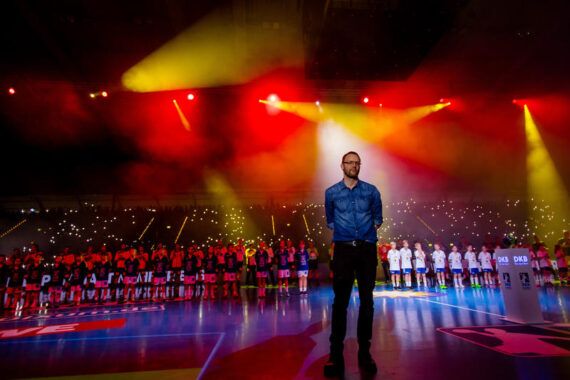 Ein Mann steht im Scheinwerferlicht einer Sporthalle, hinter ihm sind die Mannschaften aufgereiht, bunte Lichter und Rauch erfüllen die Arena. Im Hintergrund leuchten die Telefonlampen der Zuschauer.