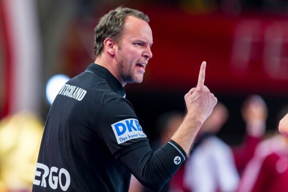 Ein Mann in einem schwarzen Hemd gestikuliert mit seiner rechten Hand und hebt den Zeigefinger, während er zu sprechen oder zu schreien scheint, was darauf hindeutet, dass er während eines Sportereignisses leidenschaftlich Anweisungen gibt oder reagiert.