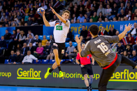 Ein Handballspieler in einem weißen Trikot springt in die Luft, um den Ball zu werfen, während ein Torwart in einem grauen Trikot sich darauf vorbereitet, den Ball zu blocken. Ein Schiedsrichter und ein dicht gedrängtes Publikum beobachten das Geschehen im Hintergrund.