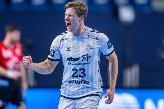 Ein männlicher Handballspieler in einem weißen Trikot mit der Zahl 23 reckt während eines Spiels die Faust in die Höhe und jubelt, während im Hintergrund unscharfe Mannschaftskameraden und Zuschauer zu sehen sind.