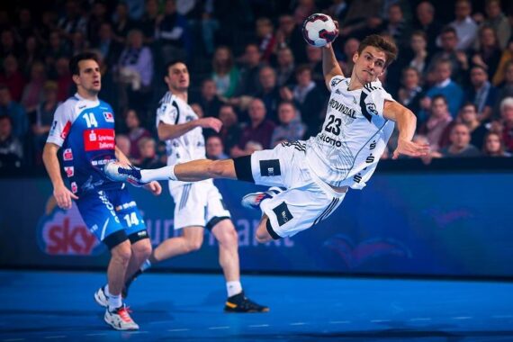 Ein Handballspieler in einem weißen Trikot springt mit einem Ball in der Hand durch die Luft und versucht, ihn zu werfen, während Spieler in blauen und weißen Trikots zusehen. Die Aktion findet auf einem Hallenplatz statt, mit einer Menschenmenge im Hintergrund.