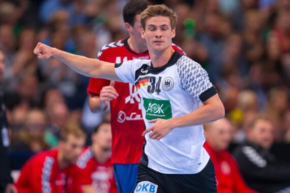 Ein männlicher Handballspieler in einem weiß-schwarzen Trikot mit der Nummer 34 ist in Bewegung und zeigt mit seinem linken Arm. Ein anderer Spieler in einem roten Trikot ist hinter ihm zu sehen. Im Hintergrund ist eine unscharfe Menschenmenge zu sehen.