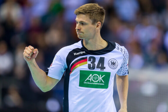 Ein männlicher Handballspieler, der ein weiß-schwarzes deutsches Trikot mit der Nummer 34 trägt, ballt während eines Spiels seine Faust in einer Geste der Entschlossenheit, während im Hintergrund eine unscharfe Menschenmenge zu sehen ist.