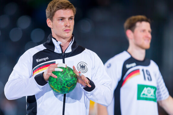 Ein männlicher Handballspieler in einer weißen deutschen Mannschaftsjacke hält einen grünen Handball, während ein anderer Spieler in einer ähnlichen Uniform unscharf im Hintergrund zu sehen ist.