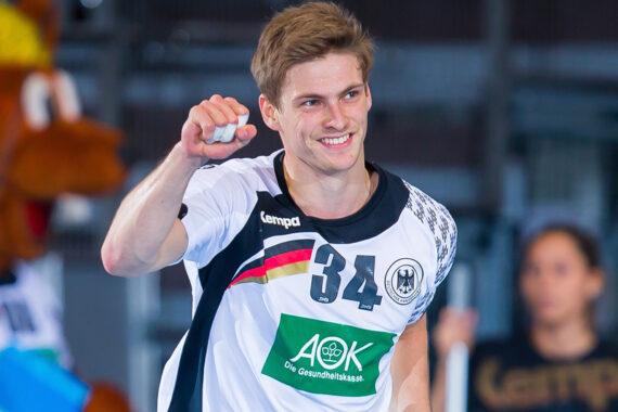 Ein männlicher Handballspieler in einem weiß-schwarzen Trikot mit der Nummer 34 lächelt und reckt die Faust zum Jubel auf einem Hallenplatz in die Höhe.