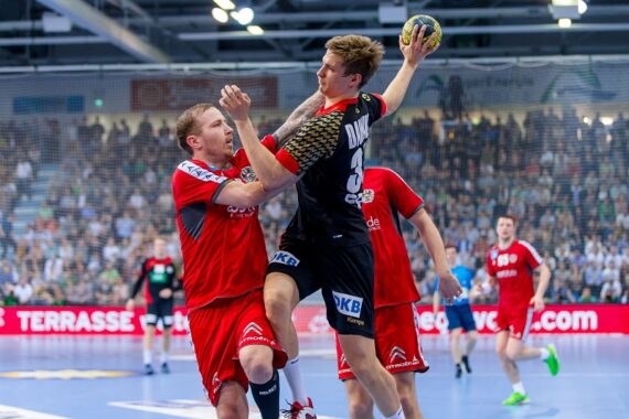 Ein Handballspieler in einem schwarzen Trikot springt, um den Ball zu werfen, während ein Verteidiger in einem roten Trikot versucht, ihn während eines Hallenspiels zu blockieren.