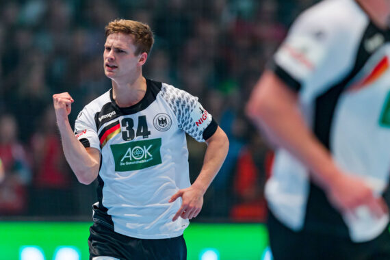 Ein männlicher Handballspieler, der ein weißes Trikot mit der Nummer 34 trägt, ballt während eines Spiels jubelnd die Faust. Im Hintergrund sind unscharfe Zuschauer und ein weiterer Spieler in ähnlicher Kleidung zu sehen.