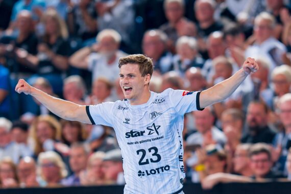 Ein Handballspieler in einem weißen Trikot mit der Nummer 23 jubelt mit erhobenen Armen und lächelnd vor einer jubelnden Menge in einer Halle.