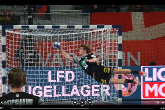 Ein Handballtorwart taucht in der Luft mit gestrecktem Arm nach links ab und versucht, einen Torschuss zu blocken. Im Hintergrund sind das Netz und einige Werbebanner zu sehen.