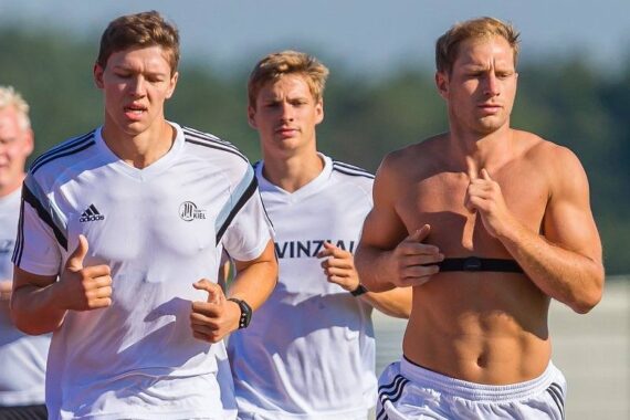 Drei junge Männer joggen im Freien; zwei tragen weiße Sporthemden, während der rechte ohne Hemd ist und ein Fitness-Tracker-Armband trägt. Die Bäume und der blaue Himmel sind im Hintergrund verschwommen.