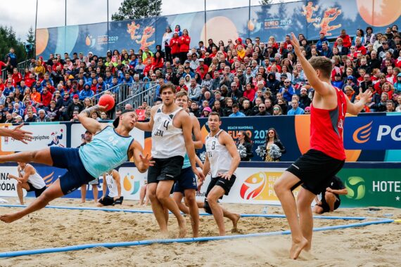 Ein Beachhandballspieler in einem hellblauen Trikot versucht einen dramatischen Flugball, während andere Spieler und eine große Menschenmenge bei einem lebhaften Spiel im Freien aufmerksam zusehen.