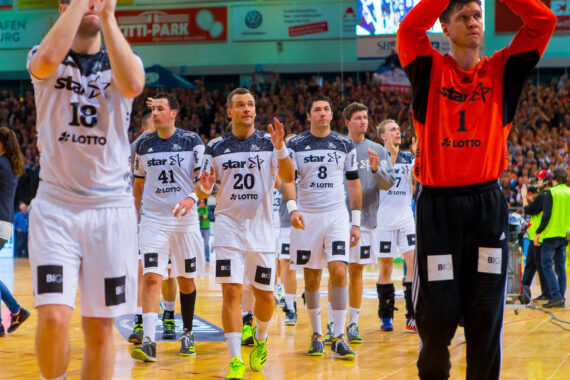 Eine Männerhandballmannschaft in weißen und schwarzen Trikots, mit einem Spieler in einem roten Trikot, läuft über einen Sportplatz und klatscht in die Hände, während die Zuschauer auf der Tribüne im Hintergrund zusehen.