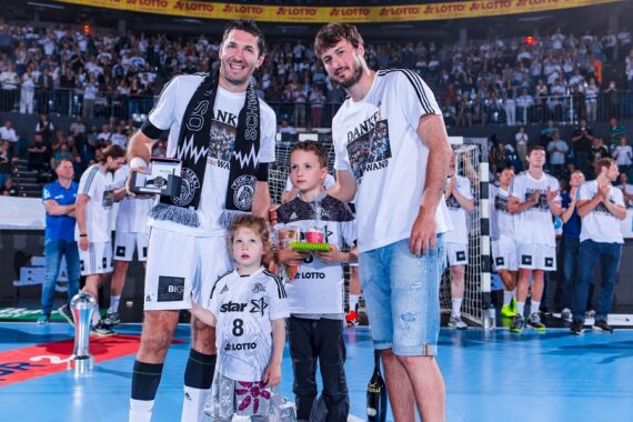Zwei lächelnde Männer und zwei kleine Kinder stehen auf einem Handballfeld und posieren für ein Foto während einer Feier. Die Männer tragen Mannschaftstrikots und Schals, und im Hintergrund ist eine Menge von Fans zu sehen.