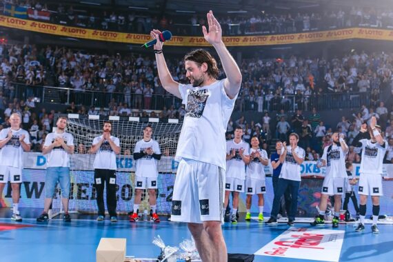 Ein männlicher Handballspieler in einem weißen Trikot steht mit erhobenen Armen und einem Mikrofon in der Hand, umgeben von applaudierenden Mannschaftskameraden auf dem Spielfeld, mit einer großen jubelnden Menge im Hintergrund.