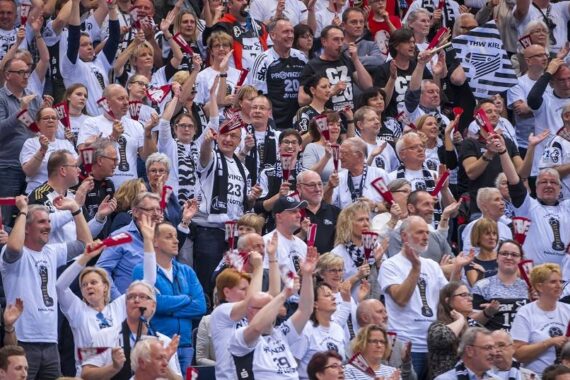 Eine große Menge begeisterter Sportfans, viele von ihnen in weißen Hemden und mit Schals, jubeln und heben ihre Arme in einem Stadion.