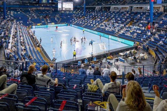 Die Zuschauer sitzen in einer großen Halle und verfolgen ein professionelles Handballspiel auf einem blauen Spielfeld. Viele Plätze sind besetzt, und die Spieler sind aktiv am Spiel beteiligt. Helle Hallenlichter erhellen die Szene.