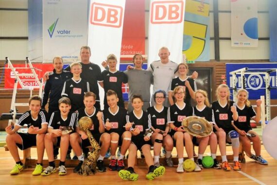 Eine Jugendsportmannschaft in schwarzen Uniformen posiert drinnen mit Trainern, die Trophäen und Medaillen halten. Sie lächeln und feiern, stehen und knien auf einem Basketballplatz mit Bannern und Sportgeräten im Hintergrund.
