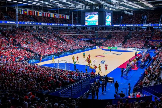 Eine große, mit Zuschauern gefüllte Halle bei einem Handballspiel während der EHF Euro 2014 in Herning. Spieler in roten und gelben Trikots treten auf einem hell erleuchteten Spielfeld an. Über den Sitzbereichen hängen Fahnen.