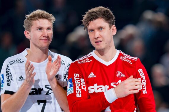 Zwei männliche Handballspieler, einer in weißer, der andere in roter Mannschaftskleidung, stehen in einer Halle nebeneinander. Der Spieler in Weiß klatscht, während der Spieler in Rot mit einem leichten Lächeln nach vorne blickt.