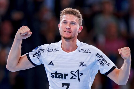 Ein männlicher Handballspieler in einem weißen Trikot mit der Nummer 7 reckt beide Fäuste in die Höhe, um auf einem Hallenplatz zu jubeln, während im Hintergrund eine unscharfe Menschenmenge zu sehen ist.