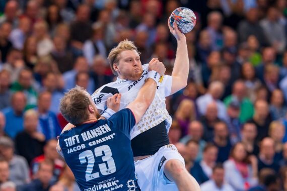 Zwei männliche Handballspieler sind während eines Spiels in Aktion. Ein Spieler in einem weißen Trikot springt mit erhobenem Ball zum Wurf, während ein anderer Spieler in einem blauen Trikot versucht, ihn zu blockieren. Im Hintergrund sieht eine Menschenmenge zu.