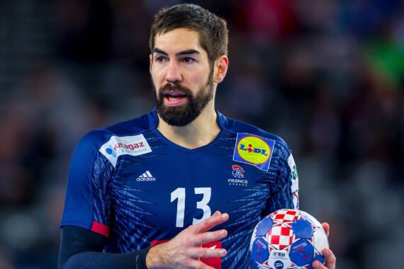 Ein männlicher Handballspieler in einem blauen Frankreich-Trikot mit der Nummer 13 hält einen bunten Handball und wirkt während eines Spiels konzentriert. Auf dem Trikot sind mehrere Sponsorenlogos zu sehen. Der Hintergrund ist unscharf.