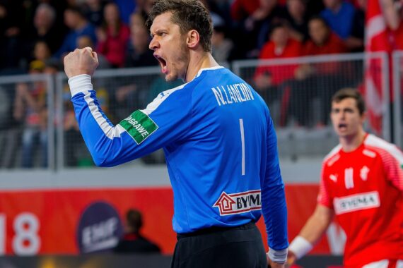 Ein männlicher Handballspieler in einem blauen Trikot mit der Nummer 1 ballt die Faust und jubelt auf dem Spielfeld, während im Hintergrund ein anderer Spieler in einem roten Trikot zu sehen ist.