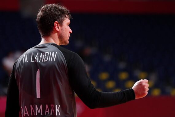 Ein männlicher Handballspieler, der ein schwarzes Trikot mit der Aufschrift N. Landin und 1 DANMARK auf dem Rücken trägt, steht mit geballter Faust und angewinkeltem Arm in einer Halle mit leeren blauen und gelben Sitzen.