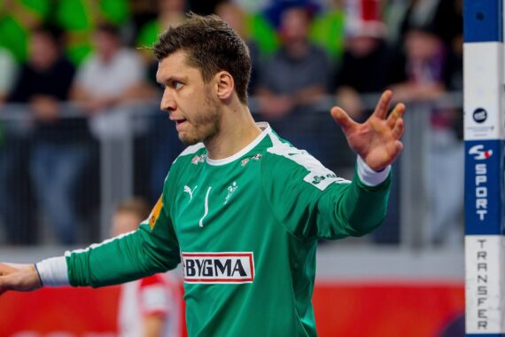 Ein männlicher Handballtorwart in einem grünen Trikot steht mit ausgebreiteten Armen, konzentriert auf das Spiel. Im Hintergrund ist eine unscharfe Menschenmenge zu sehen.