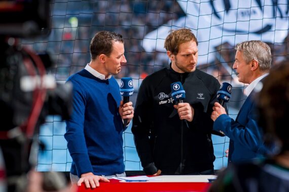 Drei Männer stehen dicht beieinander, halten Mikrofone in der Hand und führen ein Gespräch vor einem Netz in einer Sportarena. Zwei sind blau gekleidet, einer trägt eine schwarze Jacke. Fernsehkameras und helle Lichter sind zu sehen.