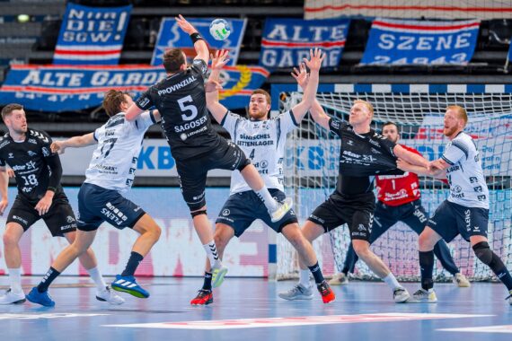 Eine Gruppe männlicher Handballspieler in schwarz-weißen Trikots spielt aktiv vor einem Tor, wobei ein Spieler springt, um den Ball zu schießen, während andere versuchen, ihn zu blockieren. Im Hintergrund hängen Transparente.