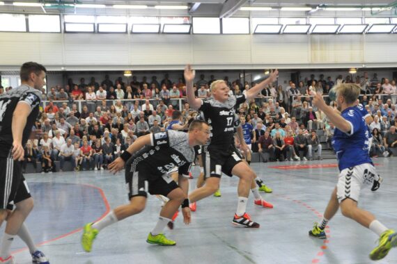 Eine Gruppe männlicher Handballspieler in schwarz-blauen Uniformen spielt aktiv in einer Sporthalle, während im Hintergrund ein großes Publikum von der Tribüne aus zusieht.