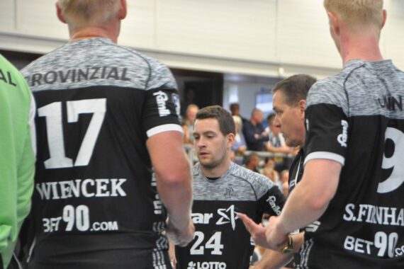 Mehrere männliche Handballspieler in schwarzen und grauen Trikots stehen zusammen auf einem Hallenplatz und scheinen eine Strategie zu besprechen. Die Szene ist konzentriert und intensiv, während die Zuschauer im Hintergrund verschwimmen.