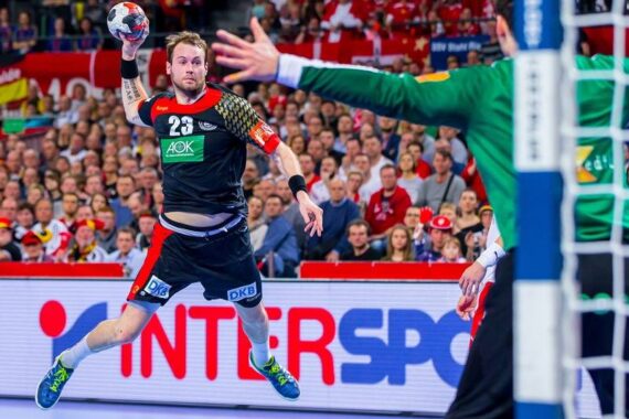 Ein Handballspieler in einem schwarzen Trikot springt in der Luft und versucht, den Ball in Richtung Tor zu werfen, während ein grün gekleideter Torwart die Arme zum Abwehren hebt. Im Hintergrund schaut eine Menschenmenge aufmerksam zu.