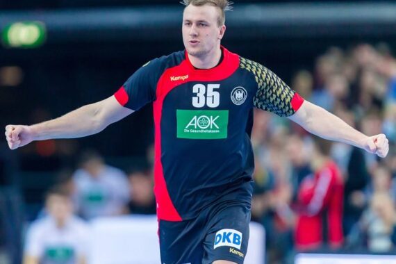 Ein männlicher Handballspieler in einem schwarz-roten Trikot mit der Nummer 35 jubelt mit ausgestreckten Armen auf einem Hallenplatz, mit einer unscharfen Menschenmenge im Hintergrund.