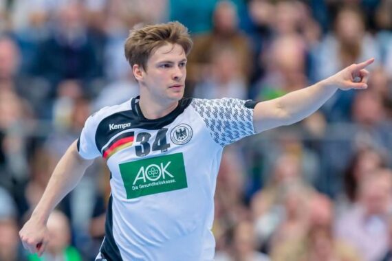 Ein männlicher Handballspieler in einem weiß-schwarzen Trikot mit der Nummer 34 und einem Streifen der deutschen Flagge zeigt während eines Spiels nach vorne. Im Hintergrund ist eine unscharfe Zuschauermenge zu sehen.