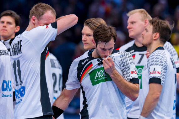 Eine Gruppe männlicher Handballspieler in weißen Trikots steht zusammen; ein Spieler küsst das Mannschaftsabzeichen auf seinem Trikot, ein anderer wischt sich mit dem Ärmel das Gesicht ab. Die Atmosphäre wirkt emotional.