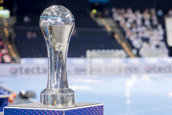 Ein glänzender silberner Pokal mit einem kugelförmigen Aufsatz steht auf einem Ständer in einer Sporthalle, im Hintergrund sind unscharfe Zuschauer und ein Handballtor zu sehen.