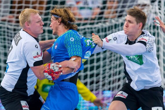 Zwei Handballspieler in weißen Trikots blockieren einen Spieler in einem blau-grünen Trikot und packen ihn an den Armen, während sie in der Nähe des Tores einen intensiven Wettkampf austragen. Im Hintergrund sind ein Netz und ein Torwart zu sehen.