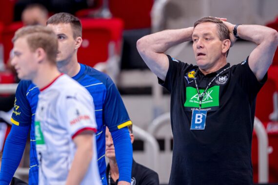 Ein Handballtrainer in einem schwarzen Hemd steht mit den Händen auf dem Kopf und sieht besorgt aus. Zwei Spieler in blau-weißen Trikots stehen im Vordergrund, und im Hintergrund sind rote Stadionsitze zu sehen.