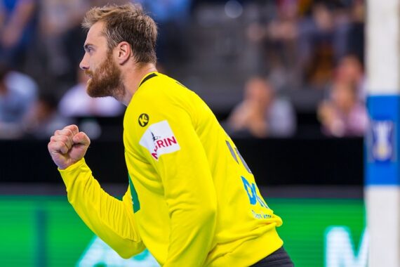 Ein männlicher Handballspieler in einem gelben Trikot jubelt mit geballter Faust und wirkt während eines Spiels begeistert und konzentriert. Der Hintergrund ist unscharf und zeigt Zuschauer und einen Teil des Spielfelds.